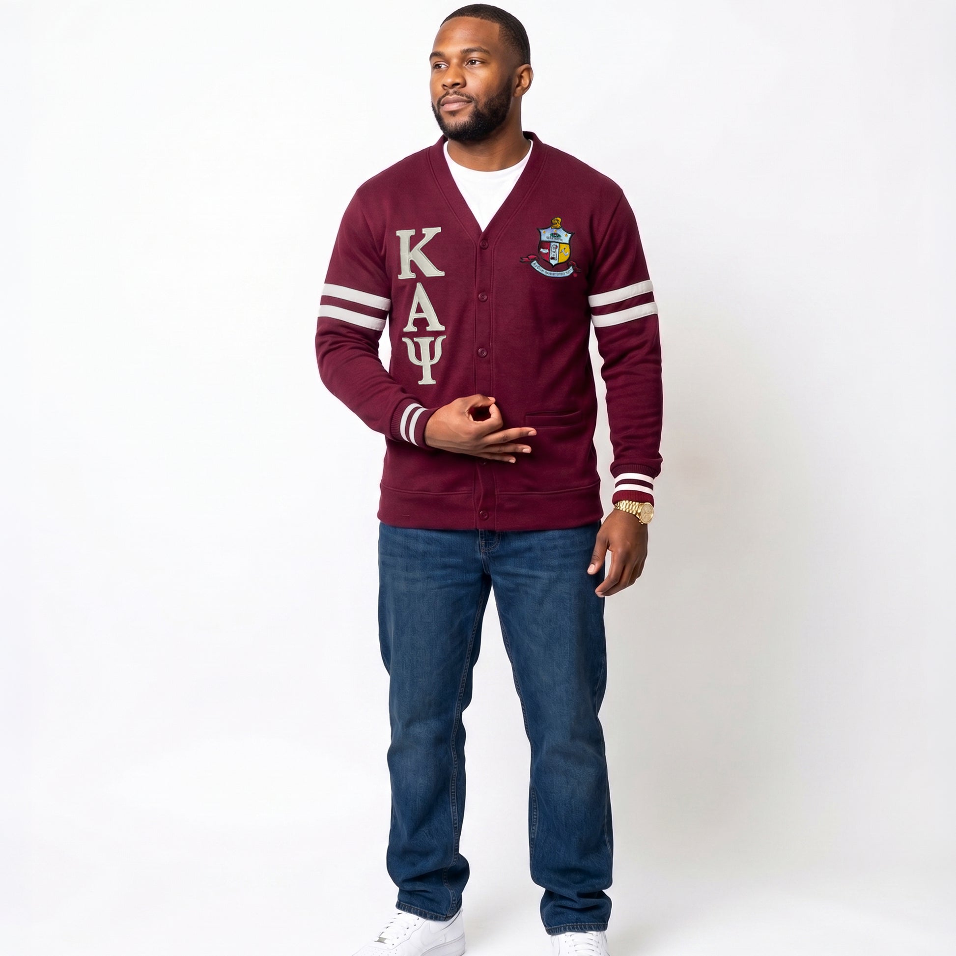 A man modeling a crimson varsity cardigan with cream lettering and stripes, paired with blue jeans and white sneakers, shot against a plain white studio background.