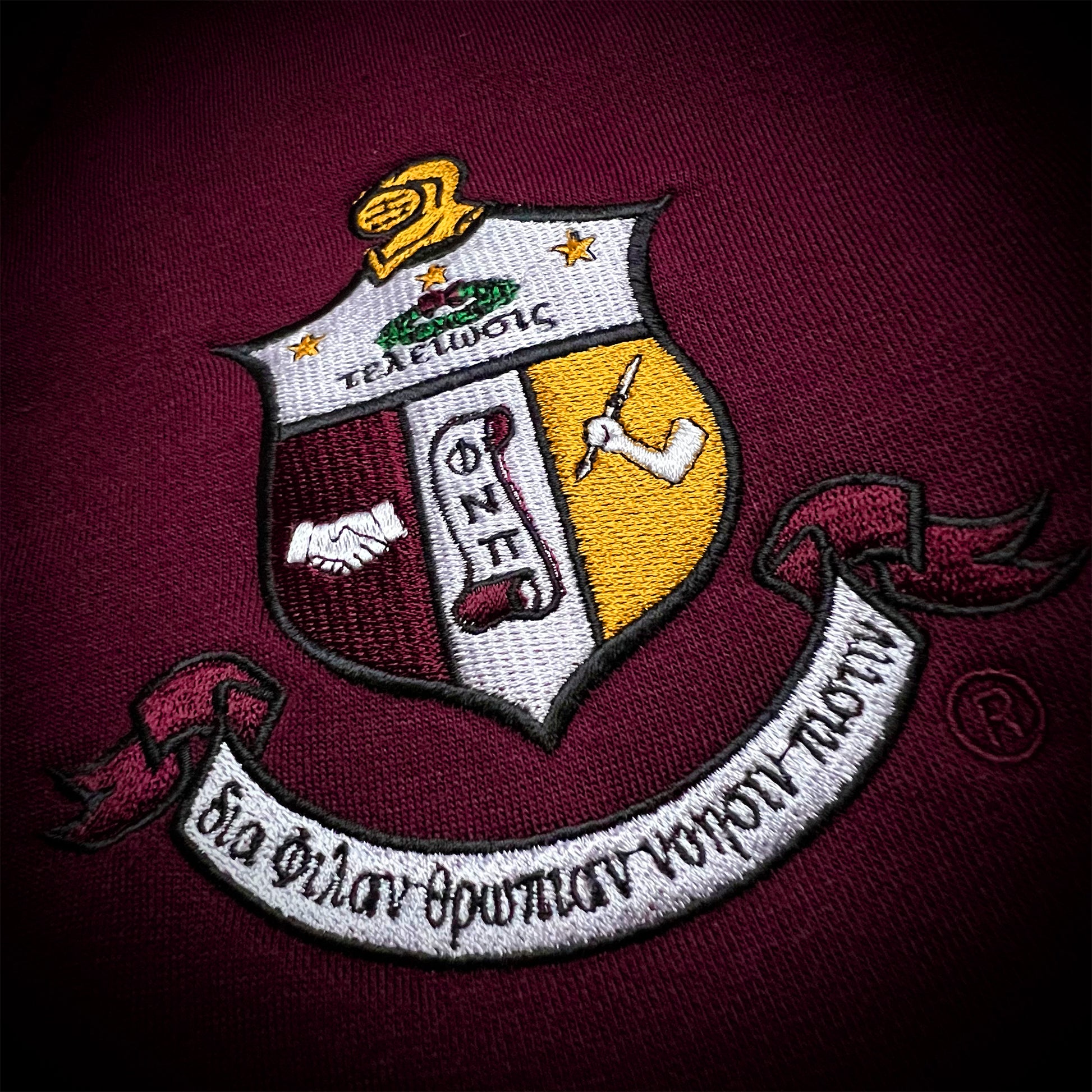 An embroidered crest patch on a crimson fabric background. The shield design is divided into white, crimson, and yellow quadrants with embroidered symbols, stars, and a white scroll banner with Greek text.